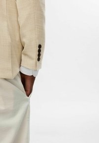 Jasnobeżowy żakiet z wzorem w jodełkę, z czterema ciemnymi guzikami na mankietach i widocznymi białymi mankietami koszuli.