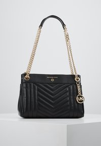 Borsa a tracolla nera trapuntata di Michael Kors con manici in catena dorata e charm con logo MK, esposta su un piedistallo bianco contro uno sfondo grigio.