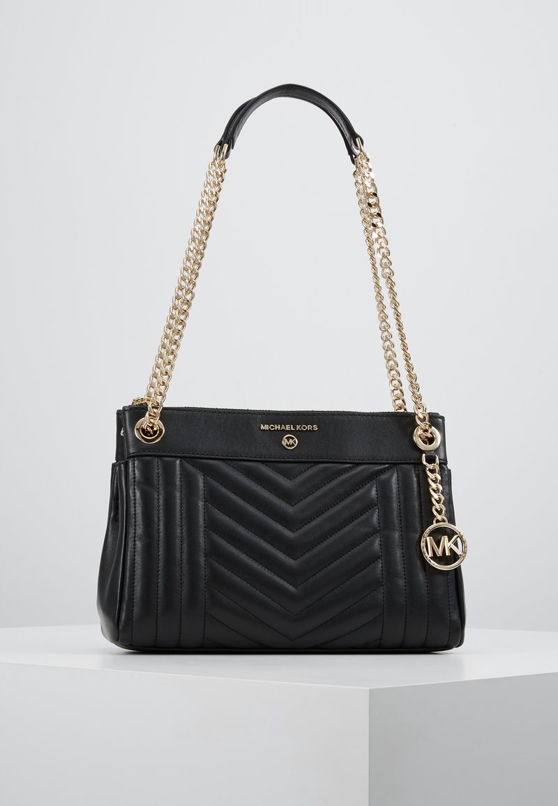 Borsa a tracolla nera trapuntata di Michael Kors con manici in catena dorata e charm con logo MK, esposta su un piedistallo bianco contro uno sfondo grigio.