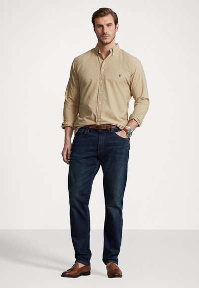 Beige skjorta med knappar, mörkblå jeans, bruna skor och armbandsur. Skjortan har en liten logotyp, en enkel design och långa ärmar.