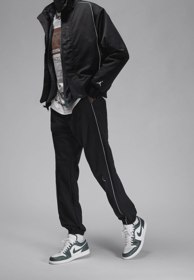 Personne portant une veste et un pantalon de survêtement noirs avec des passepoils blancs, un t-shirt graphique et des baskets blanches avec le swoosh Nike vert foncé.