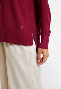 Burgunderfarbener, gerippter Strickpullover mit seitlichen Schlitzen, versehen mit einem kleinen schwarzen Logotag. Lange Ärmel mit enganliegenden Bündchen, über hellen Hosen getragen.