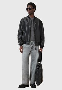 Czarna skórzana kurtka bomber, szary sweter dzianinowy, szare spodnie wide-leg, czarne buty i trzymająca czarną skórzaną torbę na ramię z suwakiem.