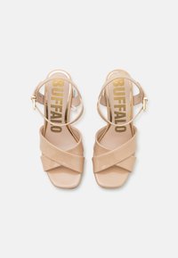 Sandalias de tacón alto de charol en color nude con tiras entrelazadas, correas de tobillo ajustables y un logotipo dorado de "BUFFALO" en las plantillas.