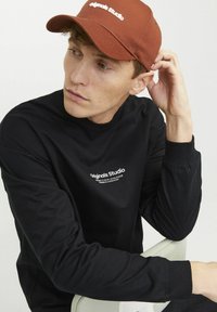 Svart långärmad skjorta med vitt tryck på bröstet och rostfärgad keps med broderad text. Slät tygkvalitet, avslappnad passform, neutral bakgrund.
