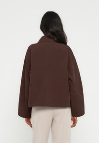 Καφέ, υπερμεγέθη μπουφάν με γιακά και χαμηλωμένους ώμους. Κατασκευασμένο από λείο ύφασμα, που παρουσιάζει μια μινιμαλιστική σχεδίαση και καθαρές γραμμές.