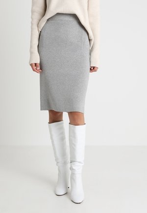 Vrouw draagt een lichtgrijze kokerrok tot op de knie, een crèmekleurige trui en witte kniehoge laarzen met hakken, staand tegen een effen achtergrond.