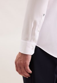 Mannen witte tekstuur lange mouwen shirtmanchet met kleine bloemen borduursel boven de pols, gedragen met donkere broeken.
