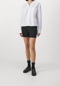 Camicia bianca con bottoni e maniche lunghe, dotata di tasca frontale, abbinata a pantaloni corti neri con bottoni laterali, indossata con mocassini neri e calze.