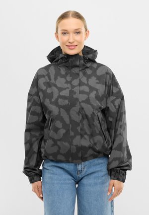 Femeie purtând o geacă cu glugă de culoare neagră și gri, cu model, manșete elastice și blugi albaștri, stând în picioare pe un fundal simplu.