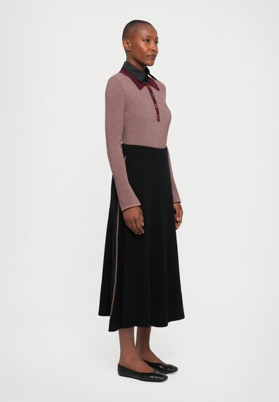 Woman standing in a mauve long-sleeve top with buttoned collar and a black midi skirt with side zipper detail and black flats.