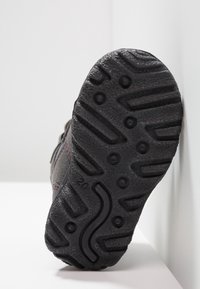 Pohled na černou podrážku boty, zobrazující detailní vzory a textury podrážky, umístěnou na světlém pozadí.