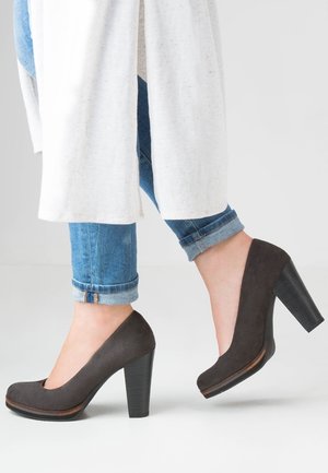 Tacones altos de ante gris oscuro con punta redondeada y tacón grueso. Los zapatos tienen un detalle de madera en la suela y se llevan con jeans remangados.