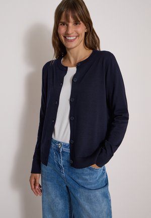 Femme souriante portant un cardigan bleu marine boutonné sur une chemise blanche et un jean bleu, debout avec une main dans sa poche devant un fond uni.
