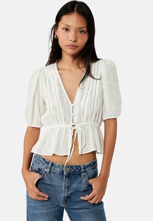 Frau mit langen dunklen Haaren, die eine weiße Bluse mit Puffärmeln und Lochstich-Details sowie blaue Jeans trägt, Hände in den Taschen, neutraler Gesichtsausdruck.