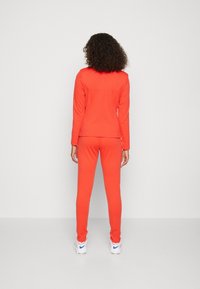 Jasnopomarańczowy, dopasowany dres wykonany z gładkiego materiału. Dopasowana bluzka z długim rękawem oraz spodnie zwężające się w kostkach. Widoczne białe sneakersy.