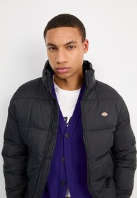 Giubbotto a sacco nero con colletto alto, chiusura zip e patch con logo. Indossato sopra un cardigan viola con bottoni e una T-shirt bianca sottostante.