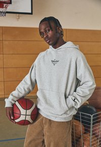 Felpa grigia con tasca frontale e logo, abbinata a pantaloni in corduroy marroni. La persona tiene in mano un pallone da basket rosso e bianco in un ambiente di palestra.