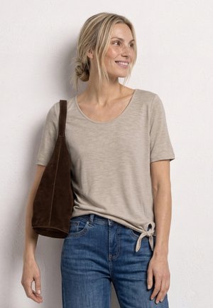 Femme aux cheveux blonds en chignon bas, portant un haut beige à manches courtes avec un lien latéral et un jean bleu, portant un sac à épaule marron foncé.