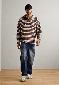 Stripete brun og hvit hettegenser med snøringer, sammen med blå jeans og grå sneakers. Modell iført svarte cap, står på tregulv.