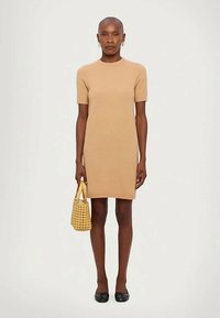 Egy bézs színű, rövid ujjú, térdhez érő ruhát viselő nő, aki világosbarna szőtt táskát tart és fekete lapos cipőt visel egy egyszínű háttér előtt.