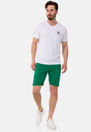 T-shirt blanc à col V et manches courtes avec logo sur la poitrine gauche, associé à un short vert jusqu'au genou. Modèle portant des baskets blanches.