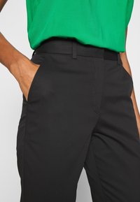 Osoba ubrana w czarne spodnie, z rękami w kieszeniach, nosząca zieloną, włożoną do środka bluzkę na tle gładkiego, jasnego tła.