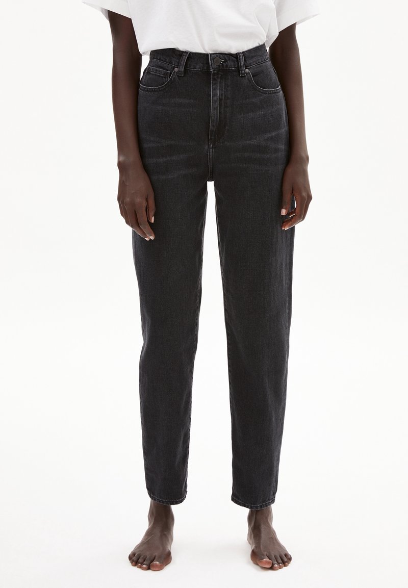 Personne portant un jean noir taille haute et une chemise blanche, debout pieds nus sur un fond blanc uni.