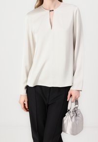 Haut à manches longues beige clair avec encolure à ouverture en forme de goutte, texture lisse, coupe décontractée, associé à un pantalon noir et un sac à main en satin argenté.