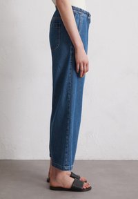 Blauwe denim broek met een ontspannen pasvorm, elastische tailleband, zijzakken en cuffs bij de enkels, gedragen met zwarte slide-sandalen.