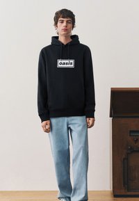 Zwarte hoodie met "oasis"-logo aan de voorkant, gecombineerd met lichtblauwe jeans met wijde pijpen. Model staat in een binnenruimte met houten meubels.