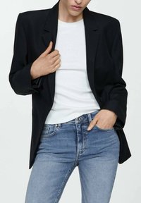 Musta bleiseri rakenteellisella suunnittelulla, yksittäinen nappikiinnitys ja viistot kaulukset, käytetty valkoisen paidan ja sinisten farkkujen päällä.