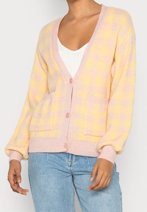 Woman wearing a pastel yellow and pink checkered cardigan with front pockets and buttons, paired with light blue jeans and a white top.