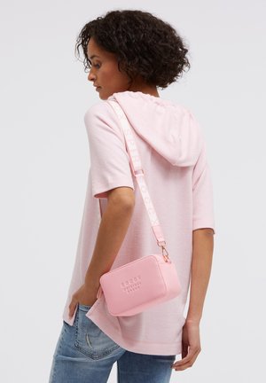 Woman in pink short-sleeve hoodie and blue jeans carries small pink crossbody bag with wide strap over her shoulder.