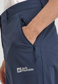 Pantaloni outdoor blu navy con tasca con zip, realizzati in tessuto liscio. Presentano un logo bianco sulla coscia. Inclusa una vita regolabile.