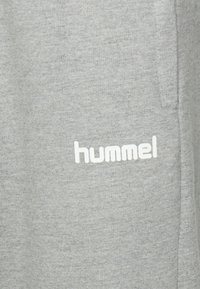 Grått tyg med den vita "hummel"-logotypen tryckt nära en sid söm och ficköppning på ett plagg.