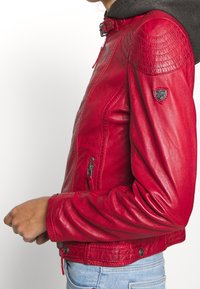 Henkilö, jolla on päällä istuva punainen nahkatakki, jonka olkapäissä on ommellut yksityiskohdat ja logopatchi, yhdistettynä vaaleansinisiin farkkuihin.