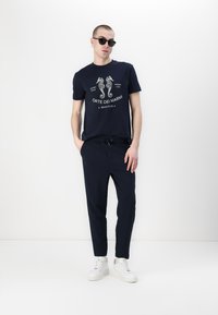 Námornícky bavlnený tričko s grafickým motívom morského koníka a logom, skombinované s tmavými jogger nohavicami a bielymi teniskami, doplnené čiernymi slnečnými okuliarmi.