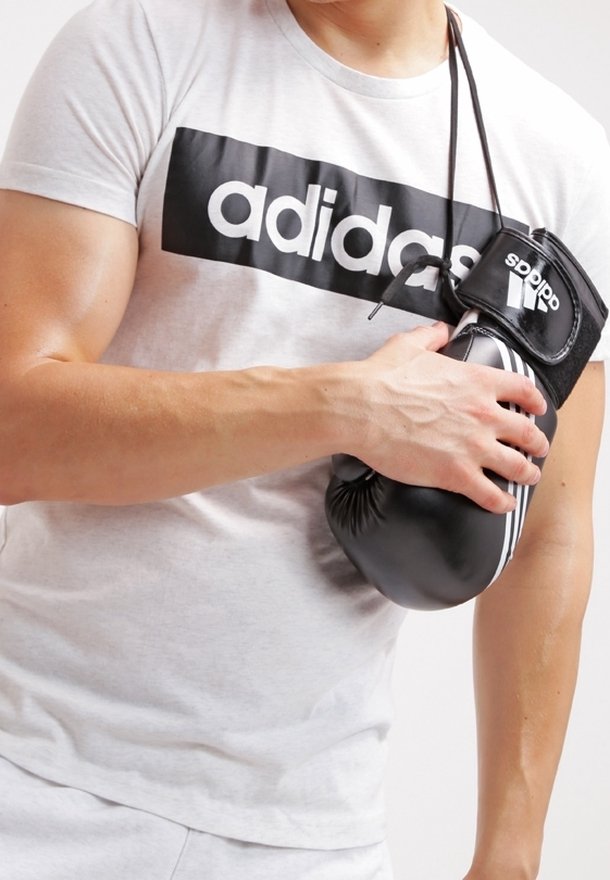 Guante de boxeo negro con rayas blancas, sostenido por una persona que lleva una camiseta gris claro con un fuerte logo negro de "adidas".