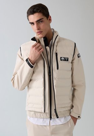 Młody mężczyzna w beżowej, warstwowej kurtce z czarnymi zamkami i logo, poprawia kołnierz na jednolitym jasnoszarym tle.