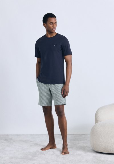 Tmavomodré bavlnené tričko s krátkymi rukávmi a malým logom v kombinácii s farebnými zelenými šortkami. Model stojí naboso na mäkkom povrchu.