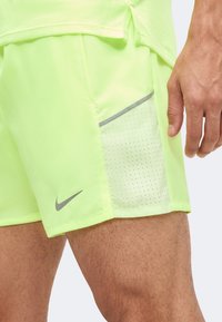 Homme portant un short de sport jaune fluo avec une poche latérale en mesh et un logo Nike swoosh gris, main reposant à côté de la cuisse.
