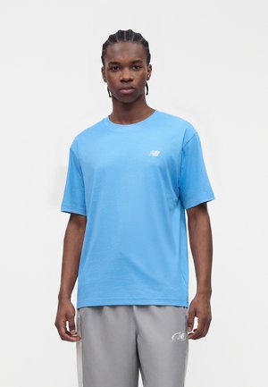 Ung mand med flettet hår iført en blå New Balance t-shirt og lysegrå New Balance bukser står mod en ensfarvet hvid baggrund.