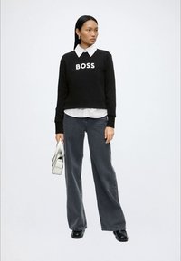 Femme portant un pull noir avec le texte "BOSS", une chemise blanche à col, un jean large foncé, des chaussures noires, tenant un sac à main blanc.