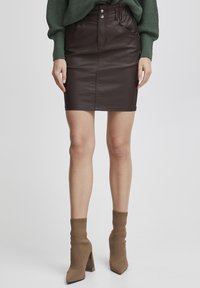 Jupe en simili cuir marron avec poches avant, fermeture à deux boutons et coupe slim. Associée à des bottines à talons beiges et des chaussettes côtelées.
