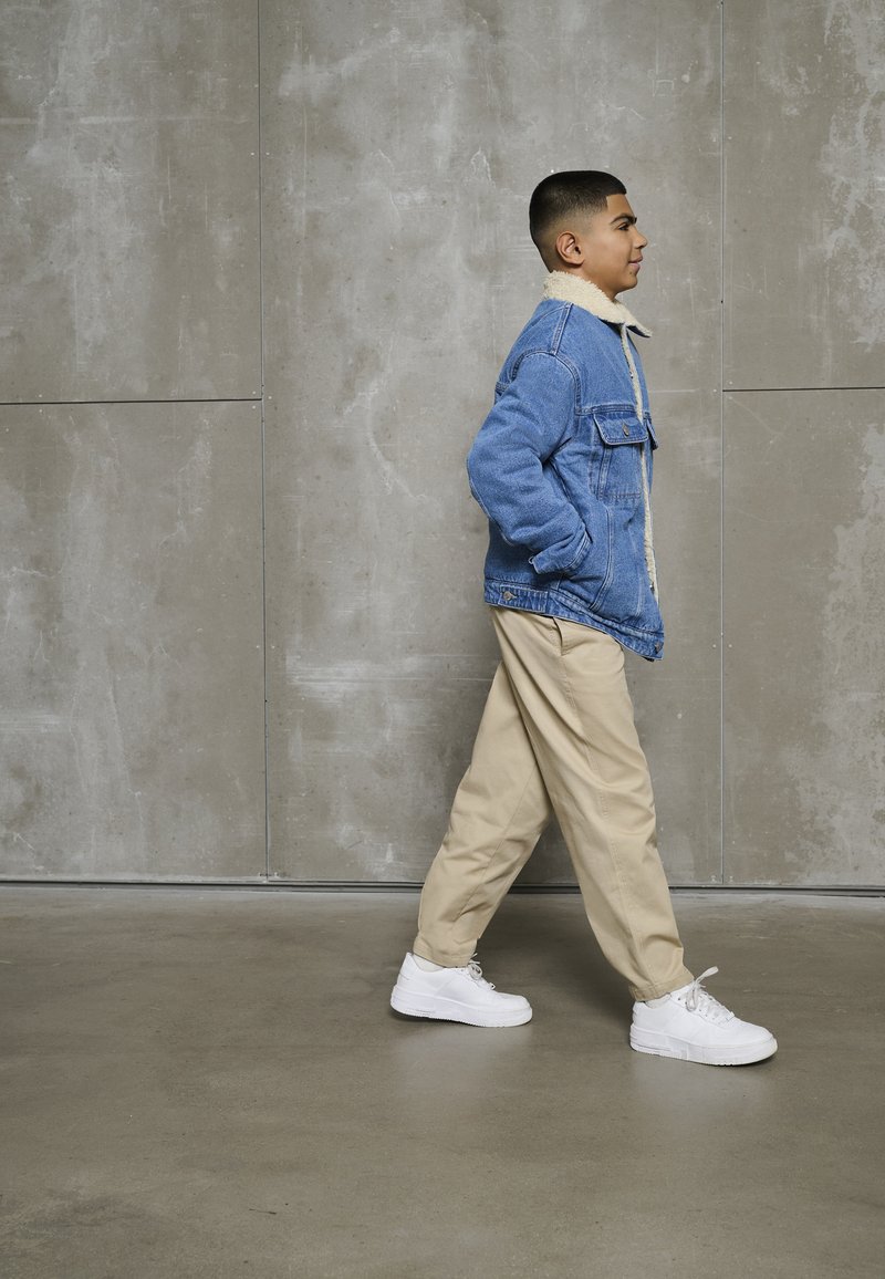 Ragazzo che cammina all'interno indossando una giacca di denim foderata in sherpa blu, pantaloni beige e sneaker bianche contro un muro di piastrelle grigio.