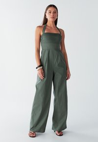 Halternek jumpsuit in een gedempte groene stof, met geribbelde accenten, wijd uitlopende pijpen en een gladde textuur. Draagt eenvoudige schoenen.