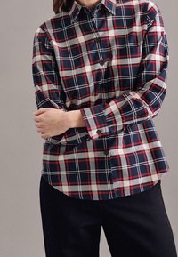 Navy, red, and gray plaid button-up shirt with a collar, long sleeves, and a curved hem; features black buttons and a chest pocket.