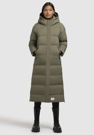 Manteau long rembourré en vert olive avec capuche, fermeture éclair à l'avant, poches latérales et couture décorative. Associé à des bottes de combat noires.