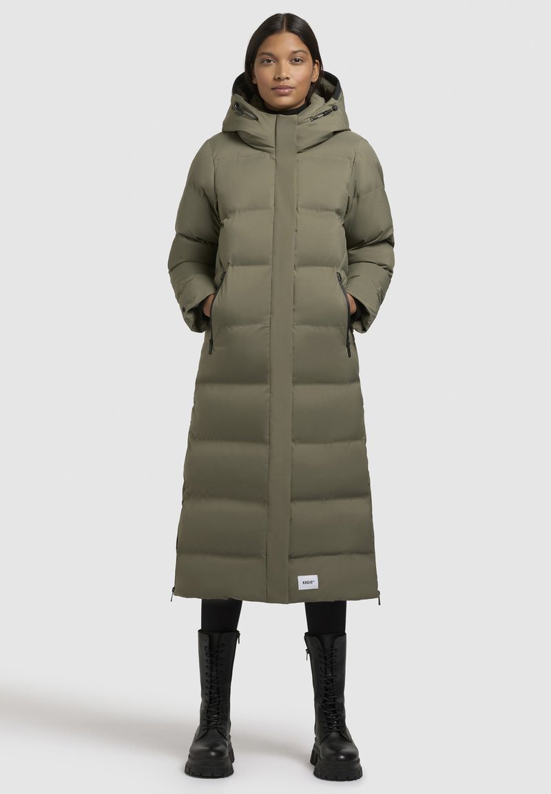 Gevoerde lange jas in olijfgroen met een capuchon, een rits aan de voorkant, zijzakken en accentstiksels. Gemaakt in combinatie met zwarte combat boots.